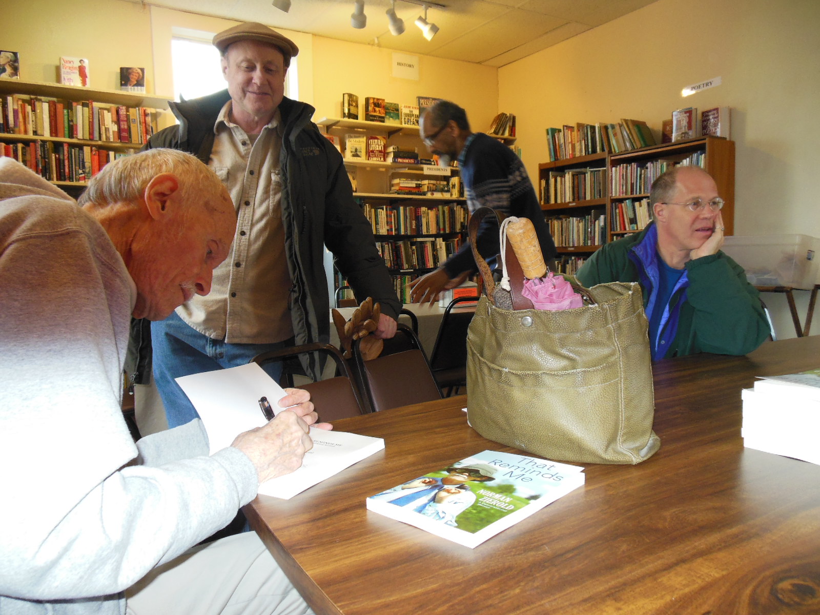 Norman Zierold, author of “That Reminds Me,” autographs one of his books for Peter Ecob Saturday at Revelations Café, after a book discussion. Freddy Fonseca, center, pushes in a chair after attending Zierold’s interview while Terry Weiss, seated, talks with others across the table.